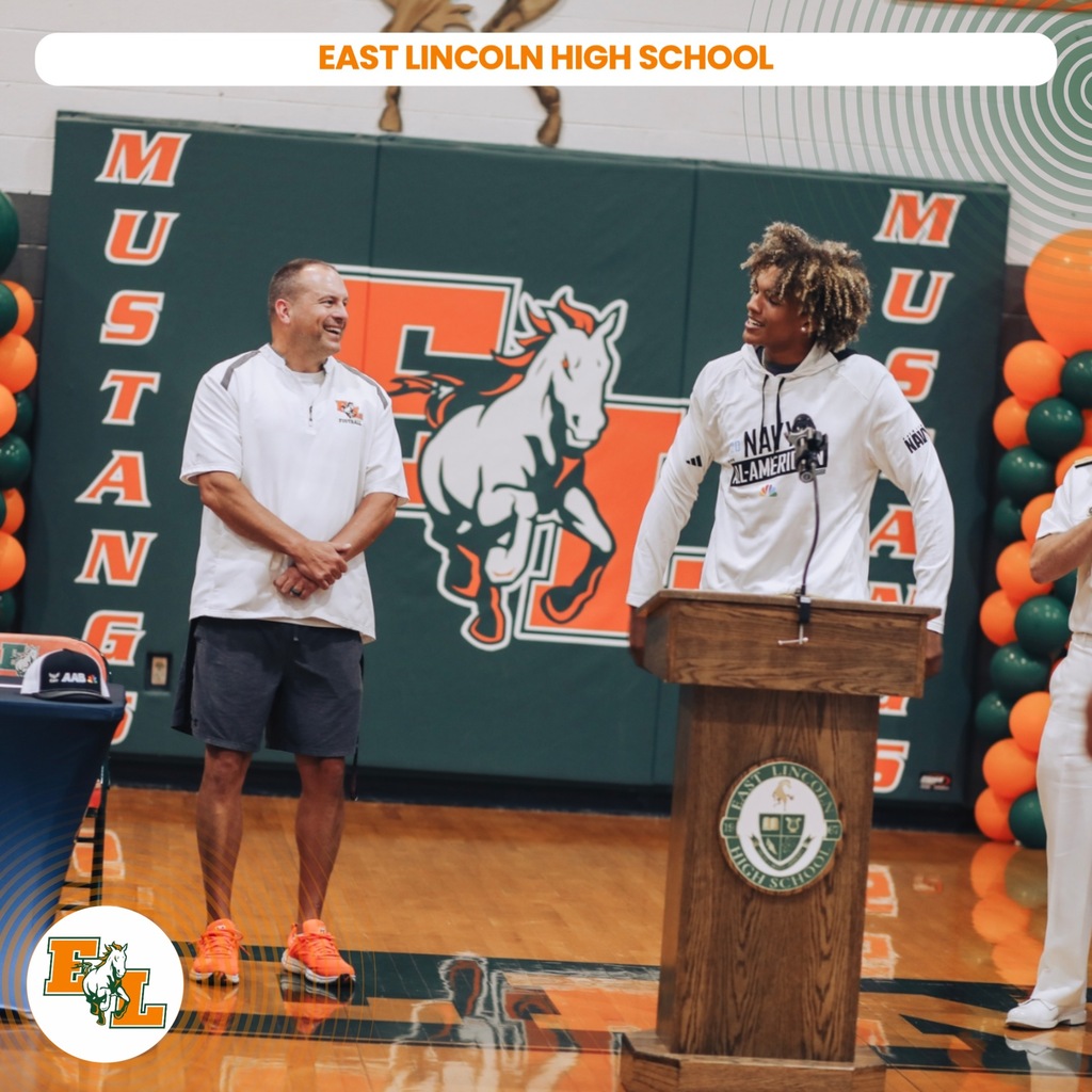 Two men on a basketball court, one speaking at a podium with a microphone, another listening.