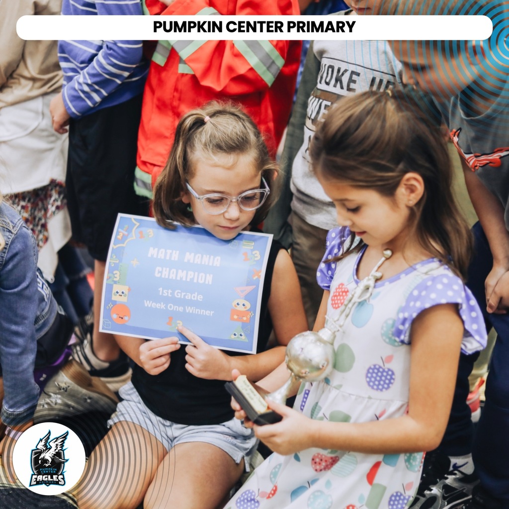 Two girls hold a blue certificate and a silver trophy. They sit in a crowd of children.