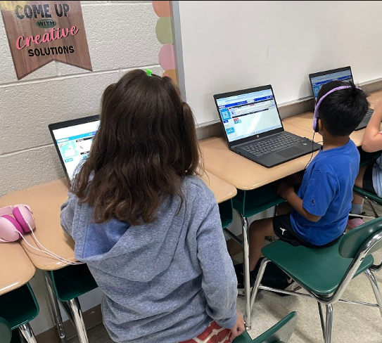 Students with headphones in a classroom setting using laptops at desks. A banner reads "Come Up Creative Solutions".
