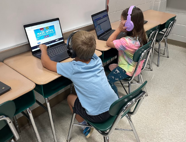 Two kids using laptops in a classroom. One is a boy in a blue shirt, and the other is a girl in a tie-dye shirt.