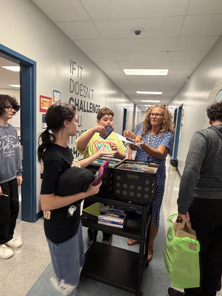 Five individuals are gathered in a hallway. A woman holds a book and talks to another woman.