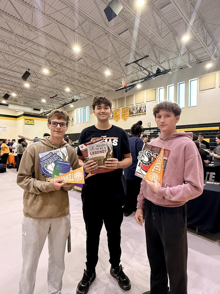 Three people in a hall, holding books. One wears glasses and a hoodie, another in a black shirt, and the third in a pink hoodie. Background shows a large hall with tables and people.