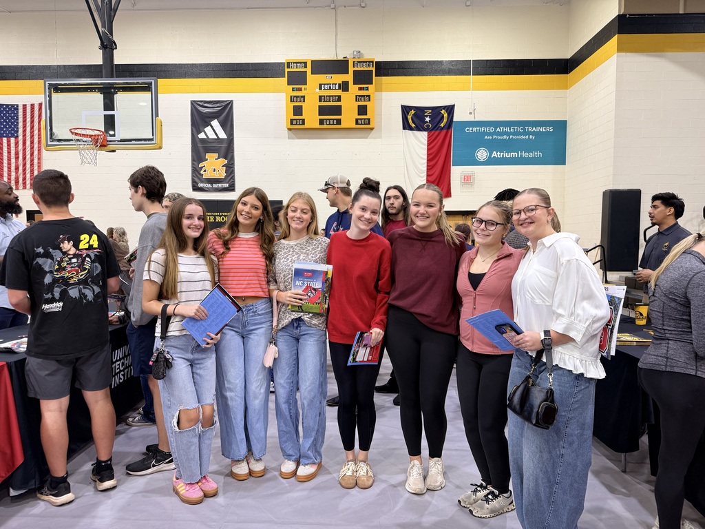Several young adults stand together in a gymnasium, holding folders and papers. They are near tables with products.