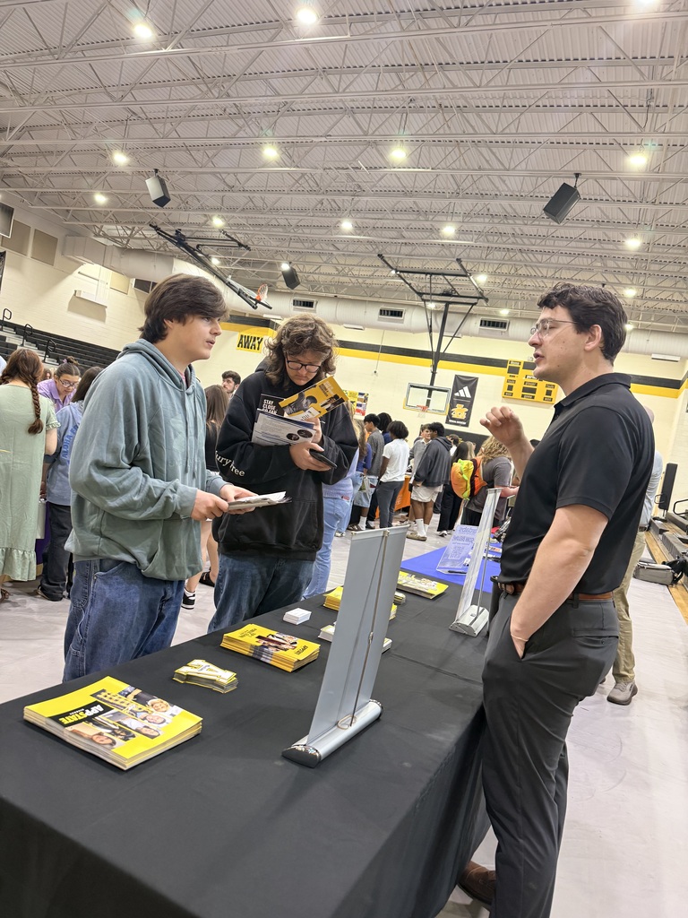 Three people stand by a table with books inside a gym. Others move about behind them.