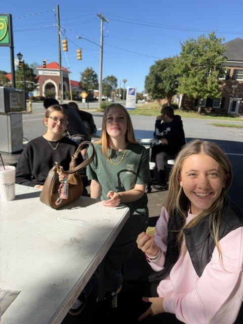 Three women are seated at a table. One holds a purse. Another holds a snack. Trees and buildings are in the background.