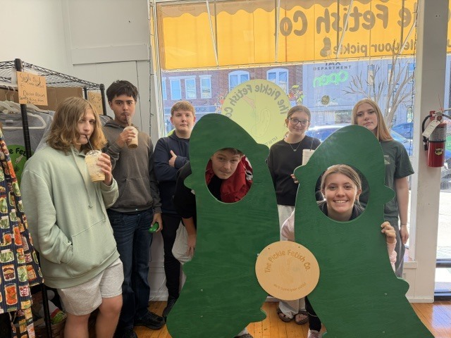 Several people stand in a room, posing for a photo. A green cutout with holes and a sign saying "Eat Fresh" is behind them.