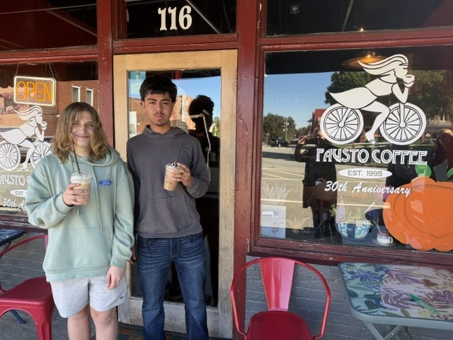 Two people stand in front of a store. The man holds a cup. The woman wears a green hoodie.
