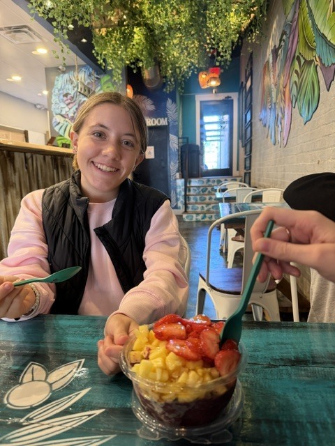 A woman sits at a table with a bowl of mixed fruit. She holds a fork. Bright plants and murals adorn the background.