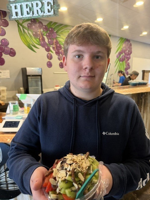 Man in dark blue hoodie holds a salad bowl with green vegetables and nuts in a kitchen.