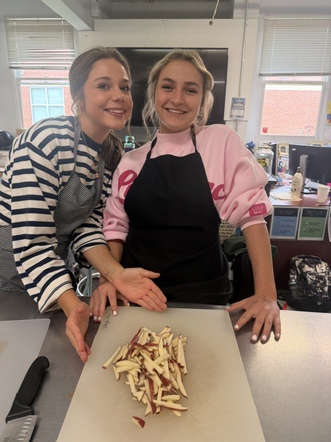 Two women in aprons prepare food on a cutting board, one holding a knife. They stand behind a table.