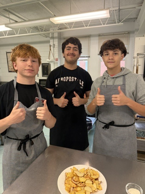 Three young men with aprons stand in a kitchen, giving thumbs-up. A plate of food is on the table.