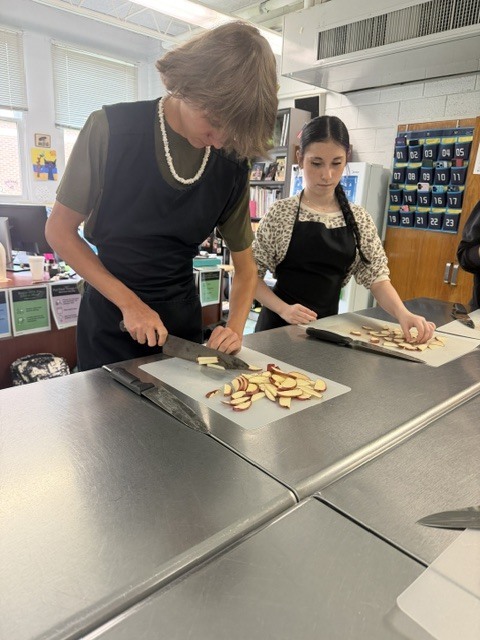 Two people wearing black aprons are preparing food on a counter in a kitchen. One is cutting.