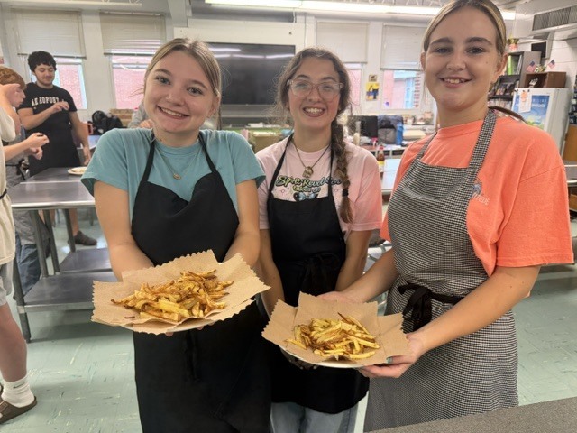 Three women in aprons hold plates of food in a kitchen. They are smiling. In the background, two other people are present.