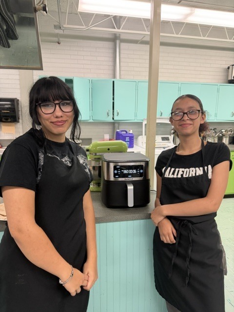 Two women wearing black aprons stand in a kitchen with teal cabinets and a black appliance.