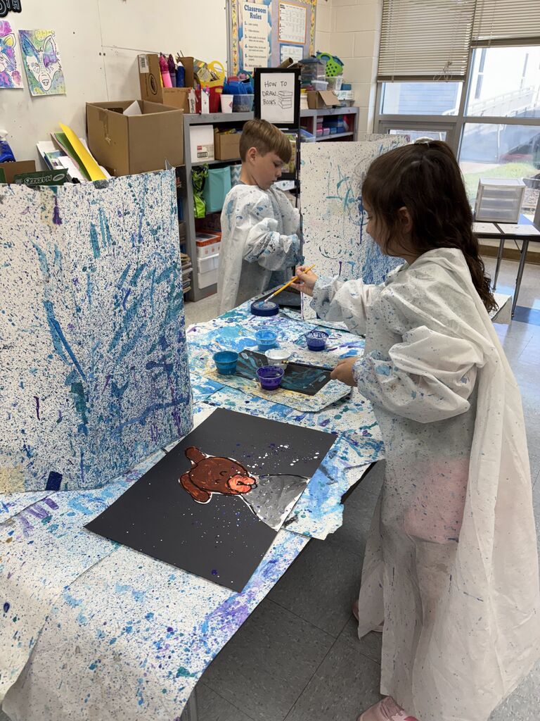 Two students wearing protective white art smocks stand at a classroom table painting individual artwork at stations separated by paint-splattered tri-fold dividers. The girl in the foreground paints a brown animal on black paper using a thin paintbrush, while small containers of blue paint and art supplies sit between the stations.