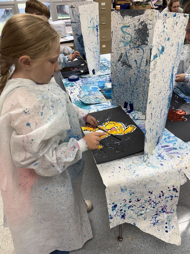 A young student wearing a protective white paint smock stands at a classroom table painting a bright yellow character on black paper. The painting station is set up with paint-splattered tablecloths and vertical dividers separating individual work areas in an art classroom setting.