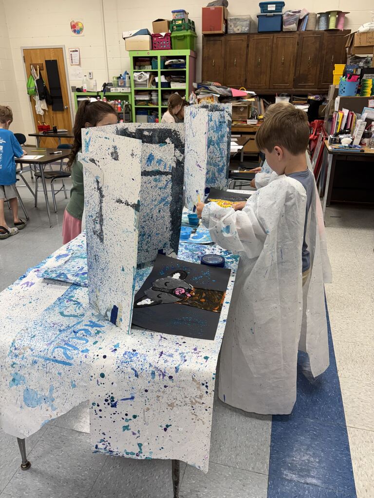 Elementary students wearing white paint smocks stand at classroom tables painting at individual stations separated by paint-splattered tri-fold boards. In the foreground, a boy on the right side of the table paints a piece of black paper while working in an art classroom filled with supplies and storage cabinets.