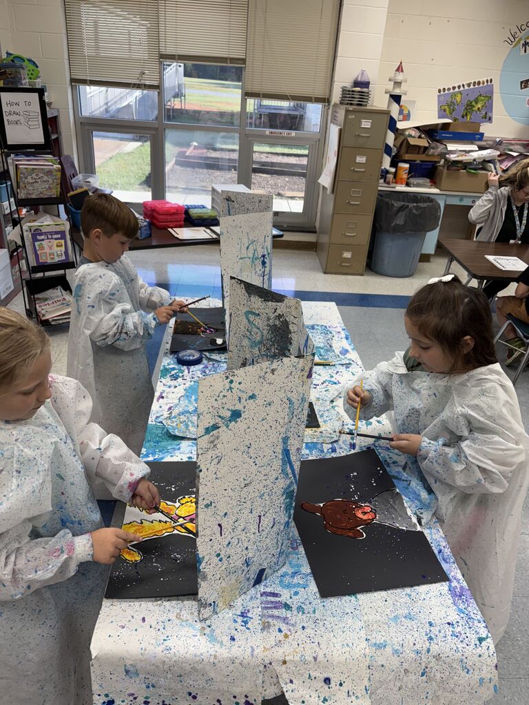 Three elementary students wearing white paint smocks stand around a table set up with paint-splattered dividers in an art classroom. The girl on the right paints a brown animal on black paper, while the two students on the left paint their own artwork at their individual stations.