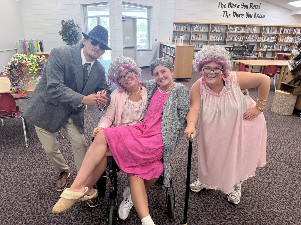 Four people dressed in old-fashioned attire pose in a library. One person sits in a chair, another stands behind, and two others stand holding canes.