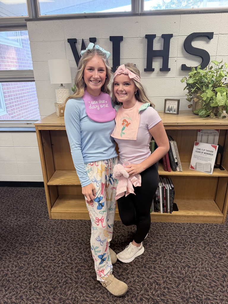 Two women in matching costumes standing in front of a shelf with books, a plant, and a lamp.