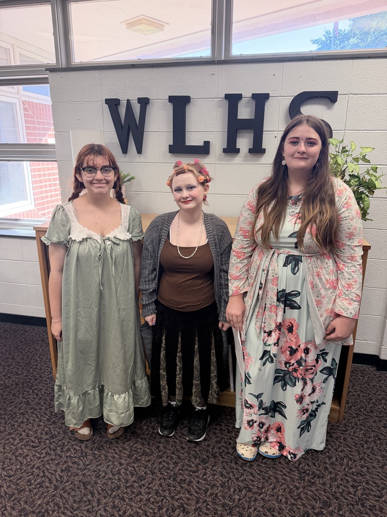 Three women in different dresses stand before a sign that reads "WLHS" on a wall.