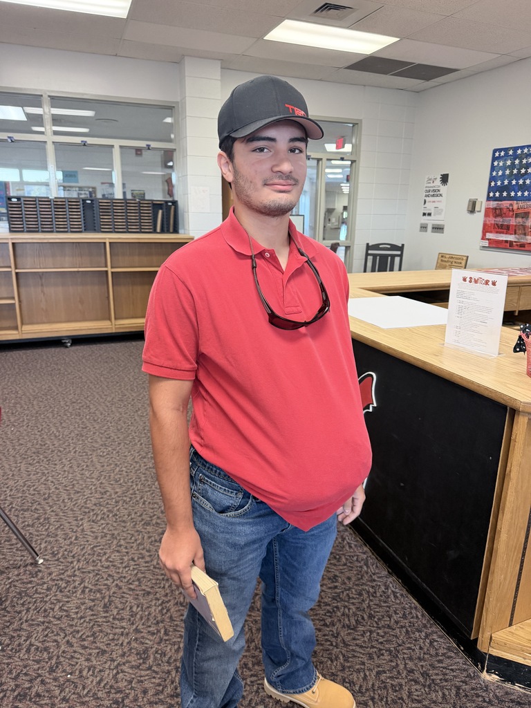 A man in a red shirt, jeans, and a cap stands in a room, holding a book. Behind him is a desk with a sign.
