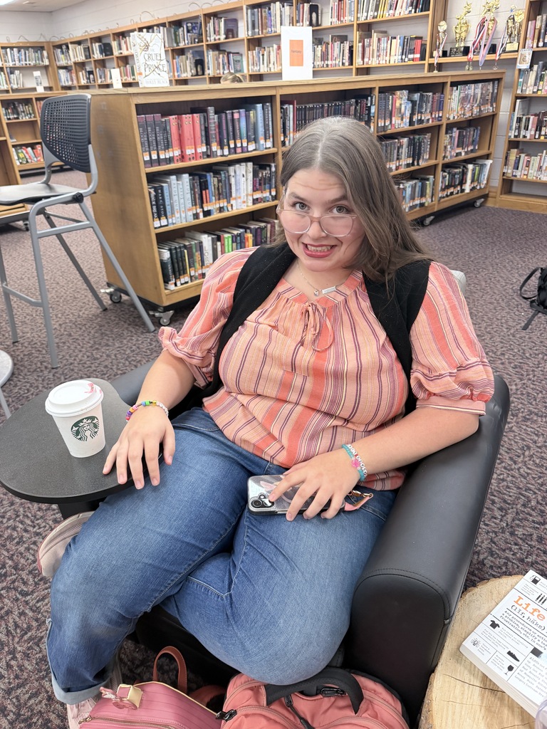 Woman sitting in a library with books in background, holding a coffee cup, smiling.