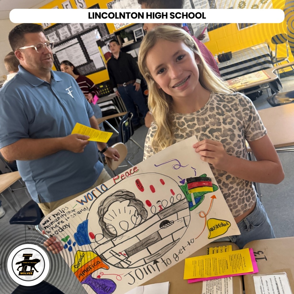 A girl with blonde hair smiles while holding up a peace poster in a classroom.