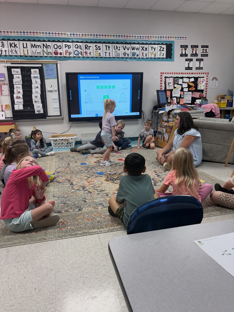 Kids sit on a rug, watching a TV screen. A woman sits on a couch. White walls with bulletin boards.