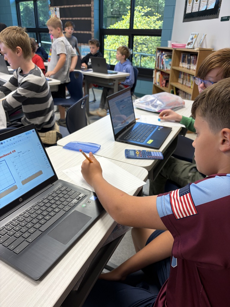 Students in a classroom with white walls and windows, working on laptops and writing. One student wears a red and blue shirt.