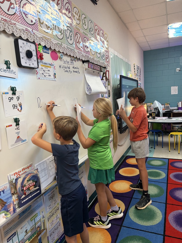 Three kids, one in green, one in blue, and one in orange, writing on a whiteboard.