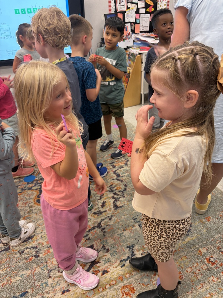 Children gather in a classroom, two girls engage in conversation while others stand behind them.