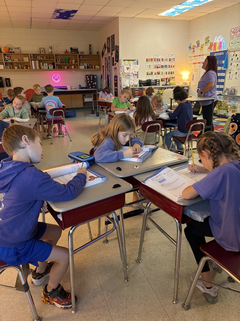 Children seated at desks in a classroom, engaged in writing, with a teacher standing at the back.
