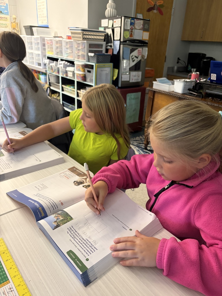 Three students, one in a green shirt, are seated at a desk and writing in their notebooks.