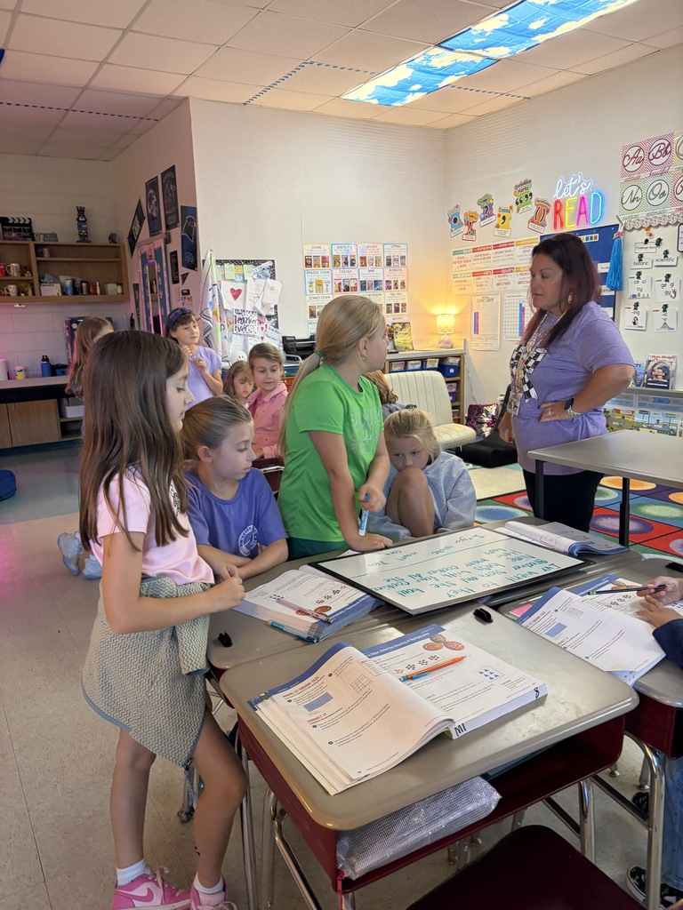 A classroom with children and adults around a table. Books and papers on the table.