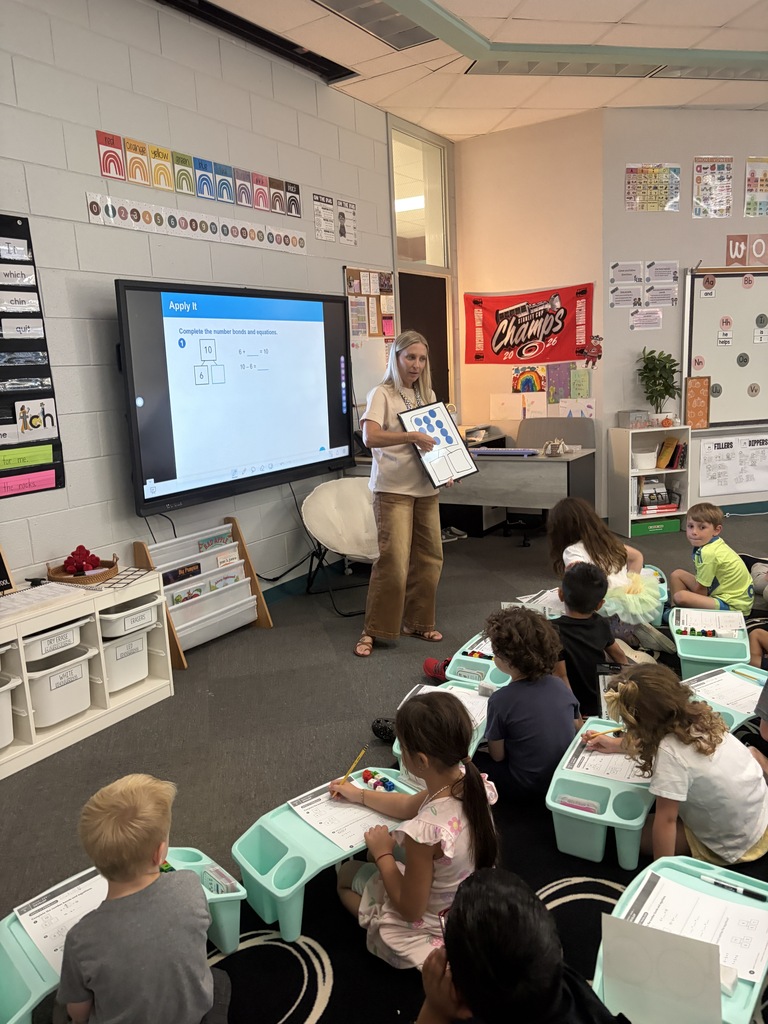 Classroom with children seated around tables, an instructor stands before a screen and holds a clipboard.