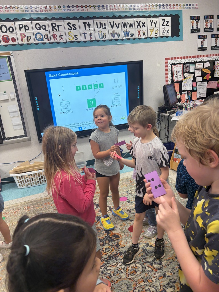 A classroom with children. Some kids are standing around while others sit on a rug. A large monitor is on the wall.
