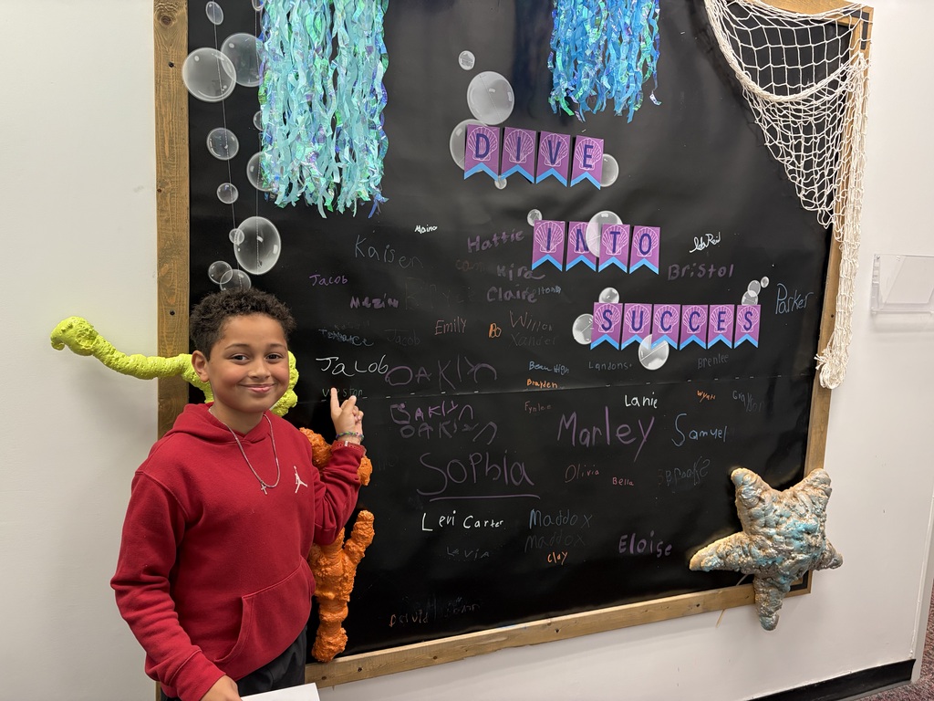 A child wearing a red hoodie smiles in front of a decorated bulletin board with blue and white designs.