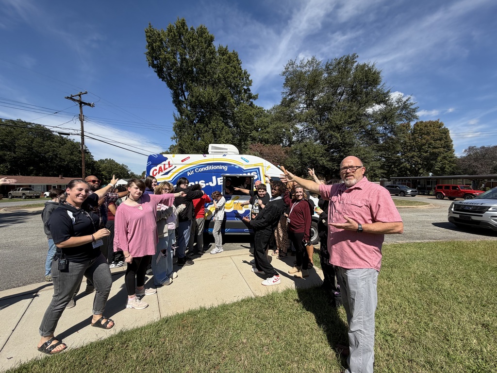 People gather by a food truck on a street. Trees and parked cars are in the background.