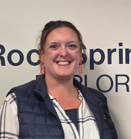 Woman smiling, standing in front of a wall with "Rock Springs" text. Wearing a blue vest over a white shirt.