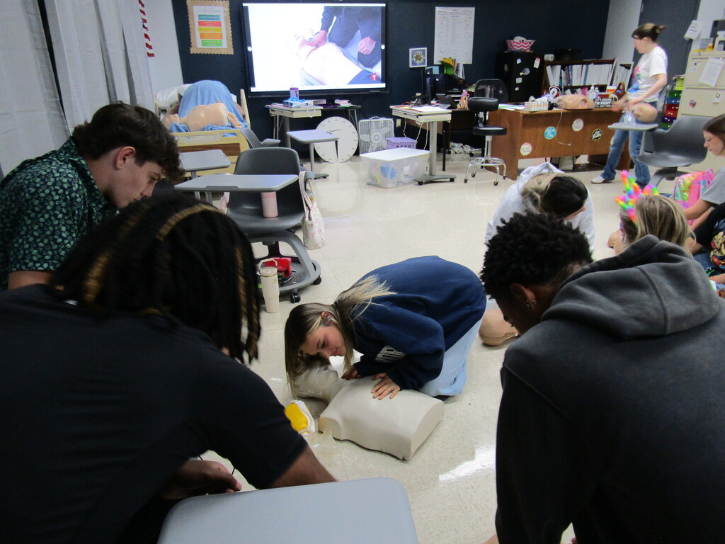 Several students practice CPR on a dummy in a classroom with desks and equipment. A person presents on a screen.