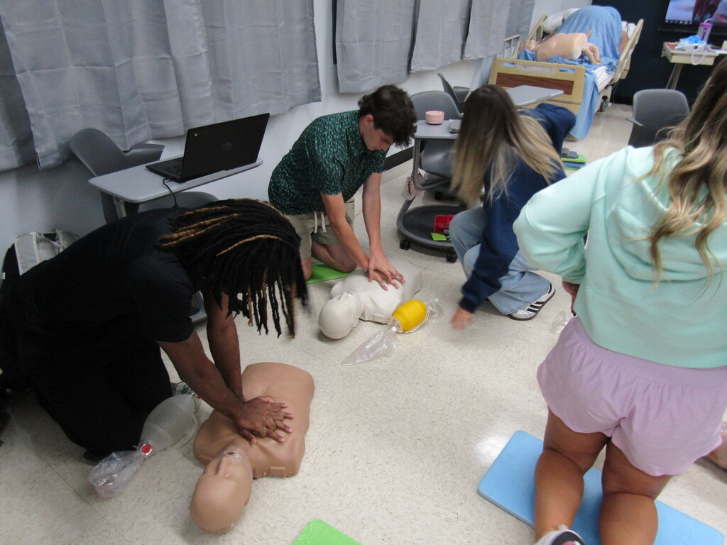 A group of people practice CPR on a dummy in a room. One person kneels, while another stands, with a desk and laptops in the background.