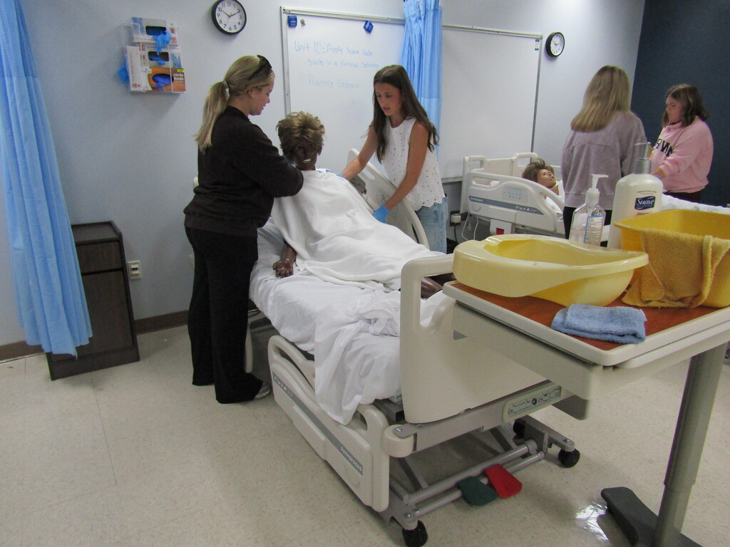 Four women are in a hospital lab; one manikin lies on a bed and another stands at the edge.