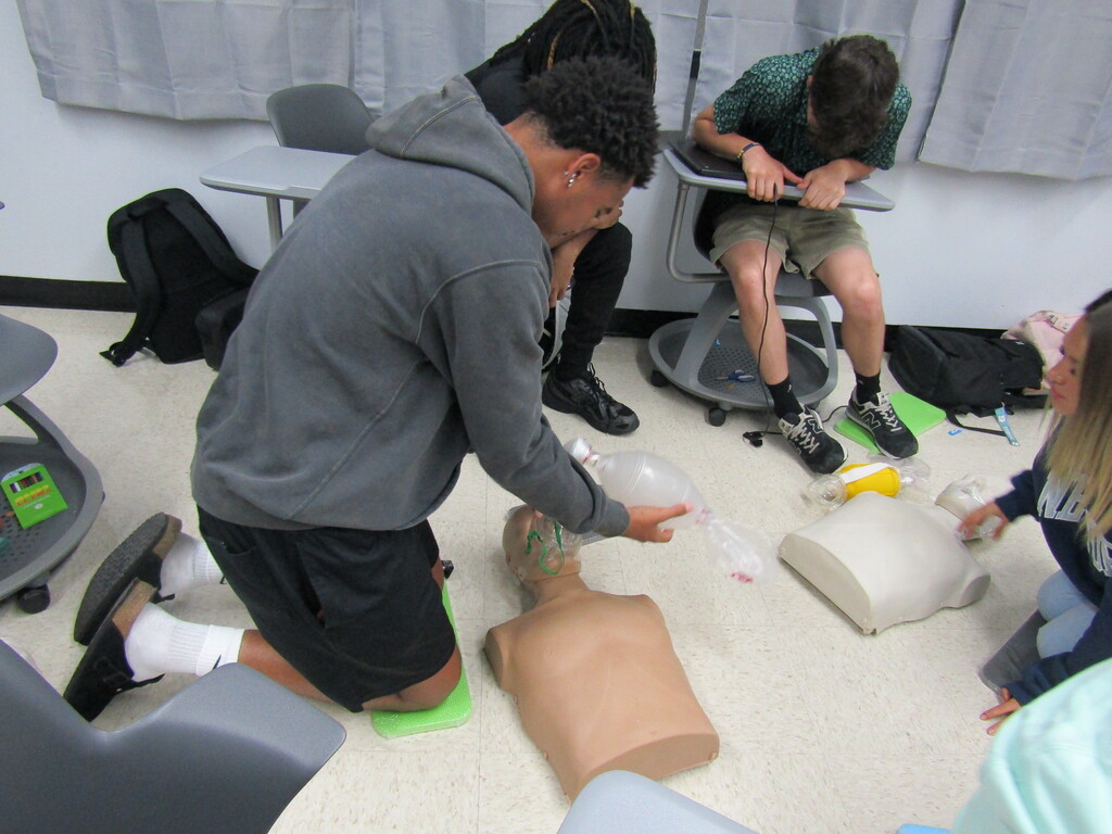 A group of people in a classroom are practicing CPR on a mannequin. Two individuals are kneeling, and others are seated.