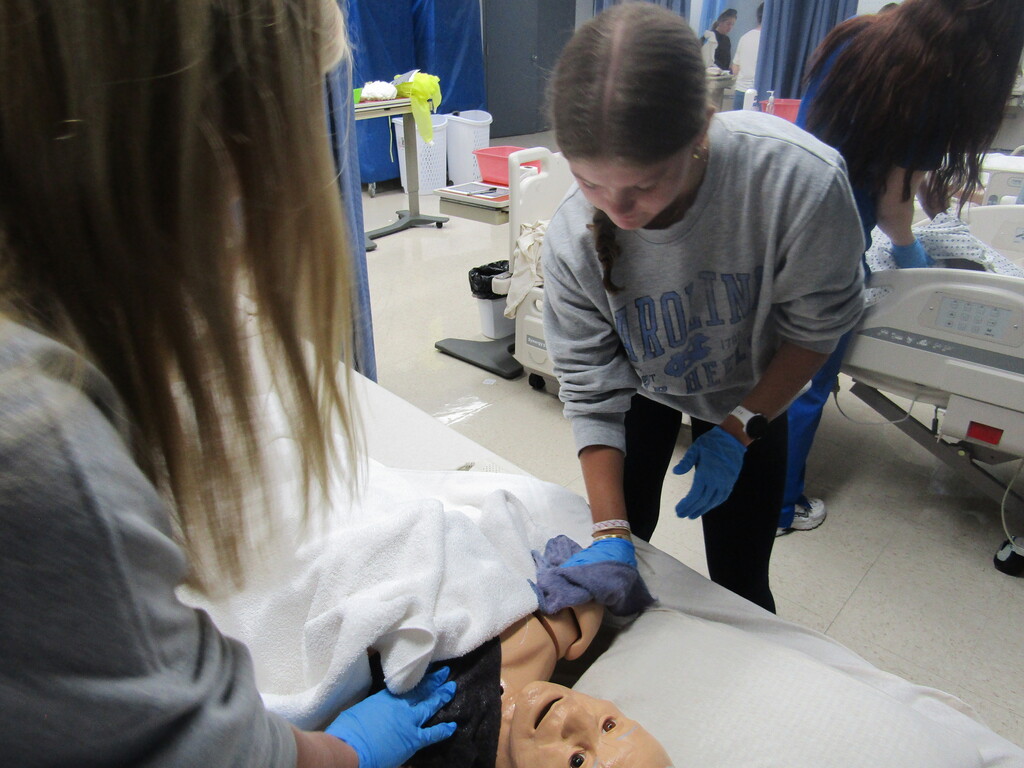 Students in a hospital practice using a mannequin, wearing blue gloves and looking at its face.
