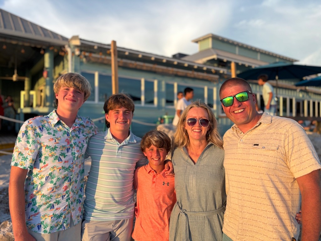 A family stands together on a beach in front of a restaurant with blue accents.
