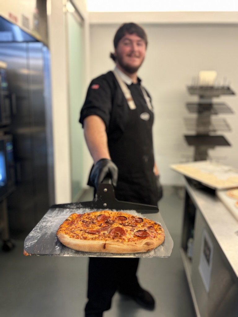 A person holds a pizza on a metal pan, with pepperoni slices. They wear a black apron and black gloves. Behind them is a counter with various items.