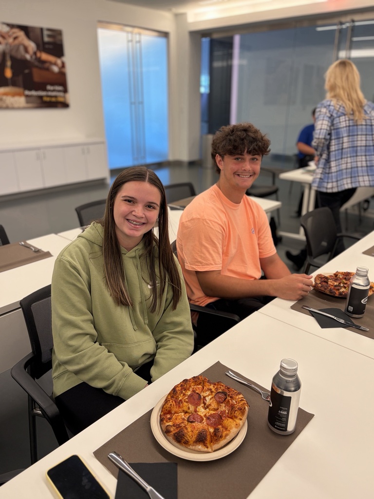 Two individuals at a table, one wearing a green hoodie and the other in an orange shirt. A pizza and drinks are on the table.