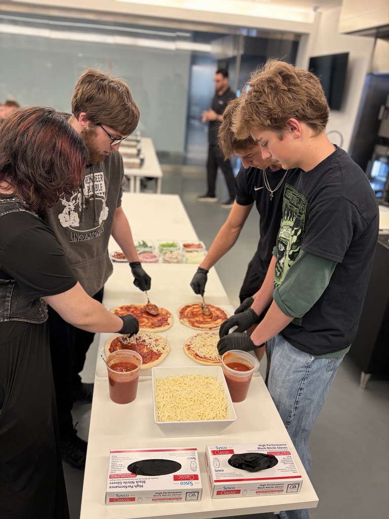 Several people, one wearing glasses, make pizzas on a table with multiple boxes of dough.
