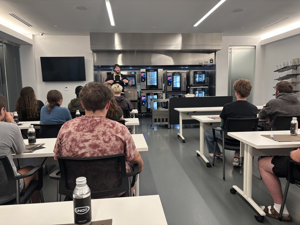 A room with white walls has people sitting at tables, facing a person standing at a counter.